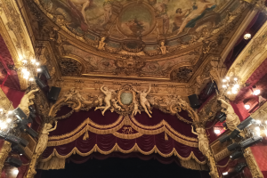 Theaterinnenraum mit roten Vorhängen, Kronleuchtern, Deckenmalerei und Skulpturen, mit einer Bühne mit einigen Menschen davor.