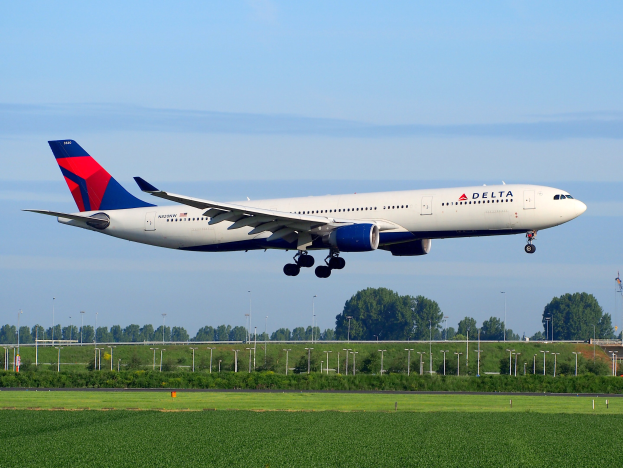 Delta Air Lines Boeing 787-9 Dreamliner beim Abheben vom Frankfurt Airport, mit grünem Gras darunter, Laternenmasten und Bäumen im Hintergrund, einem Turm auf der rechten Seite und weißen Wolken am Himmel.