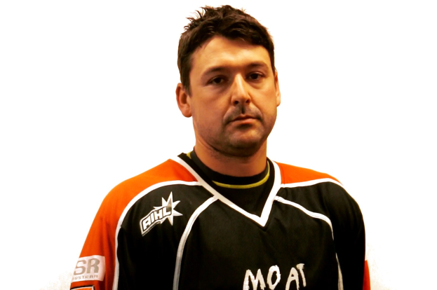 Ein Mann in einem schwarzen und orangen Hockey-Jersey mit ernster Miene, gefalteten Händen, kurzem dunklen Haar und Bartstoppeln steht vor einem weißen Hintergrund.