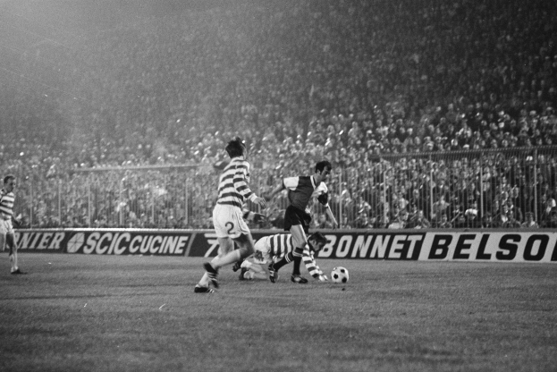 Ein schwarz-weißes Bild von Männern, die auf einem Feld Fußball spielen, mit Zuschauern auf den Tribünen und einem Banner mit der Aufschrift "Celtic v West Bromwich Albion - FA Cup Third Round."