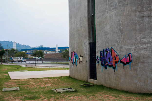 Betongebäude mit Graffiti, umgeben von Gras, Bäumen, Strommasten, Fahrzeugen auf der Straße, anderen Gebäuden, Hügeln und einem klaren blauen Himmel.