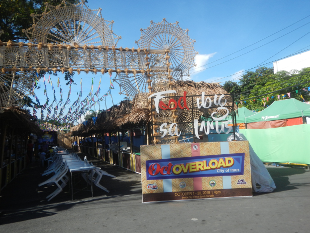 Eine Karussellszene mit einem großen Riesenrad in der Mitte, umgeben von Hütten, Tischen, Stühlen, einem Schild, Dekorationen, Bäumen, Überlandleitungen und einem bewölkten Himmel.
