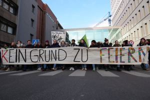 Protestierende halten ein Transparent mit der Aufschrift "Kein Grund zu Feiern" gegen deutsche Sparmaßnahmen, mit Gebäuden, einem Kran und einem bewölkten Himmel im Hintergrund.