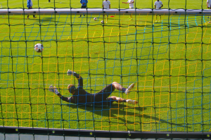 Ein Fussballspieler hält den Ball vor einem Tor, mit einem Netz im Vordergrund und einigen Menschen im Hintergrund, während mehrere Fussbälle auf dem Boden verstreut sind.
