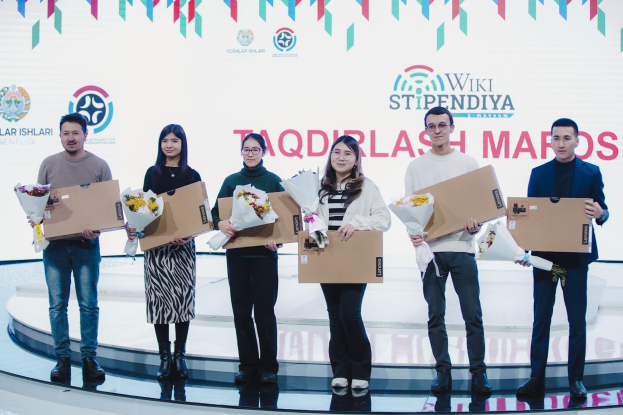 Gruppe von Menschen auf einer Bühne mit Blumensträußen und Kartons mit einem Banner im Hintergrund, auf dem "Yoslar Ishlari" und "Wikki Stipendiya" steht.