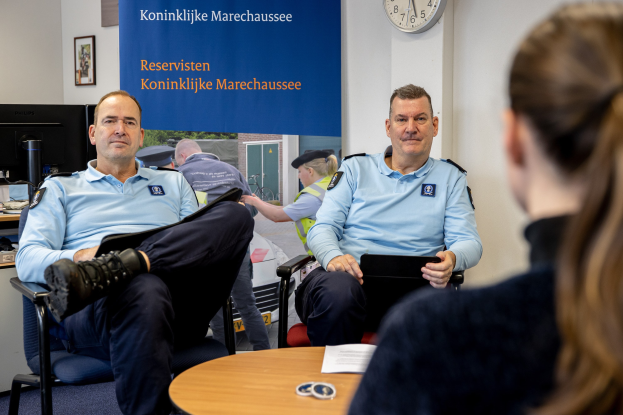 Zwei Polizeibeamte sitzen an einem Tisch mit einer Frau, umgeben von Papieren und Gegenständen, mit einer Fahne, Uhr und Foto Rahmen an der Wand und einem weiteren Tisch mit einem Monitor und Zubehör links.