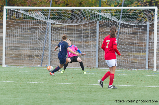 Eine Gruppe von Frauen, die auf einem grünen Rasenfeld Fußball spielen, umgeben von Bäumen, mit einem Tor im Hintergrund und Text am unteren Bildrand.