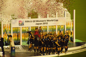Gruppe von Frauenfußballspielerinnen in schwarzen Uniformen, die auf einer Bühne feiern, umgeben von einer Menge mit Kameras, mit einem dekorierten Hintergrund und einem grasbewachsenen Boden.
