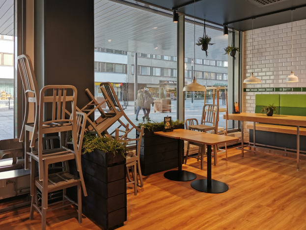Ein Restaurant mit Holztischen und -stühlen, die durch ein großes Fenster zu sehen sind, umgeben von Pflanzen und Lichtern, mit Blick auf eine Straße in Berlin mit Fußgängern, Gebäuden und Pfählen.