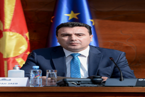 Ein Mann im Anzug und Krawatte sitzt an einem Tisch mit einem Mikrofon, Flaschen, einem Glas und einer Namensschilder, mit Flaggen im Hintergrund.