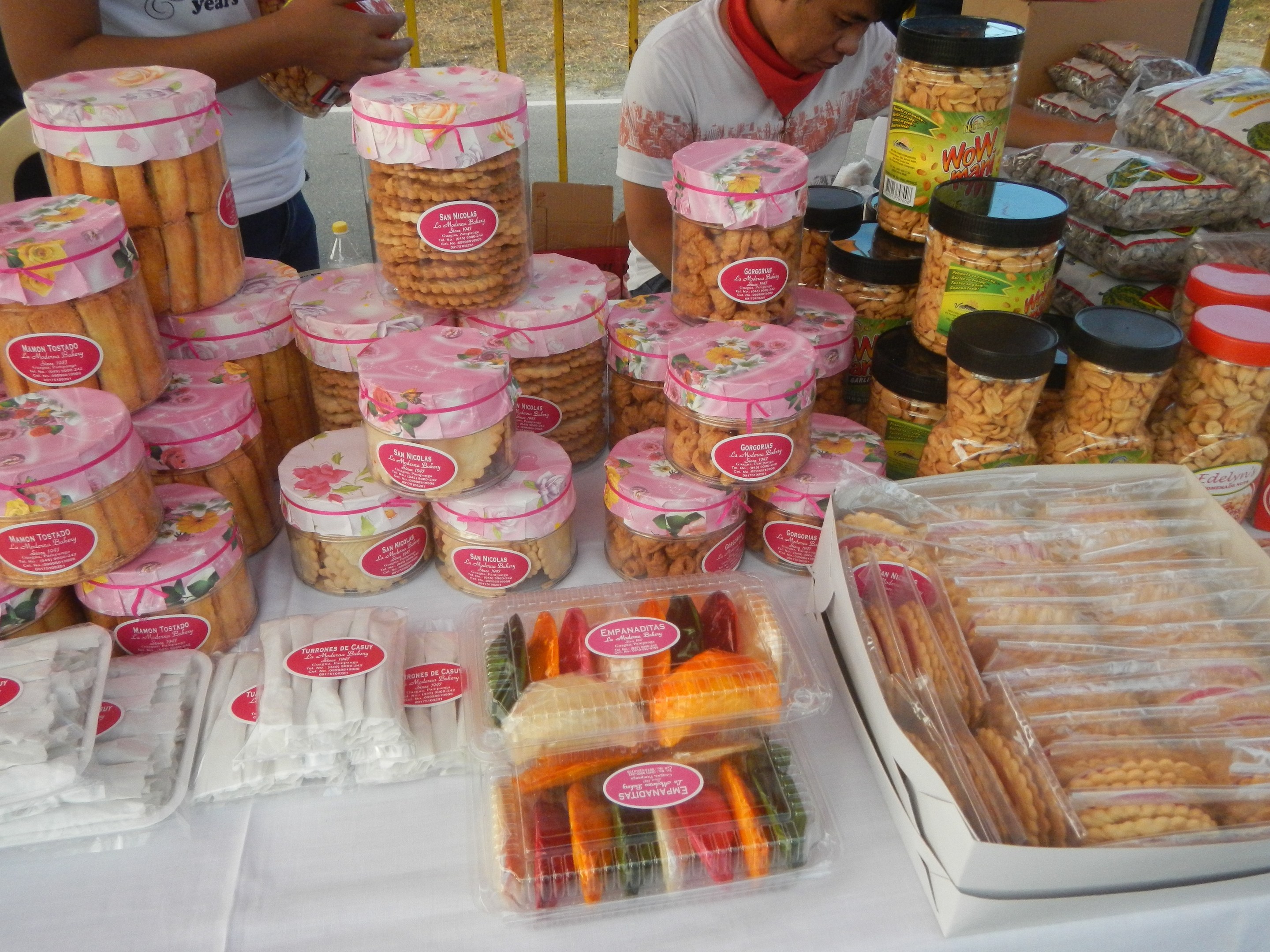 Eine Gruppe von Menschen um einen Tisch mit verschiedenen Speisen bei einem Outdoor-Food-Festival, mit einem Gel├Ąnder und einer Stra├če im Hintergrund.