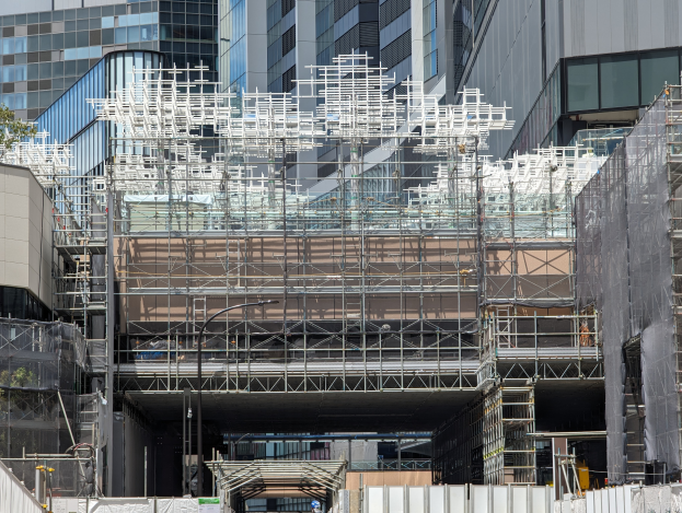 Ein großes Gebäude mit Gerüst, umgeben von Bäumen links und einer Straße unten.