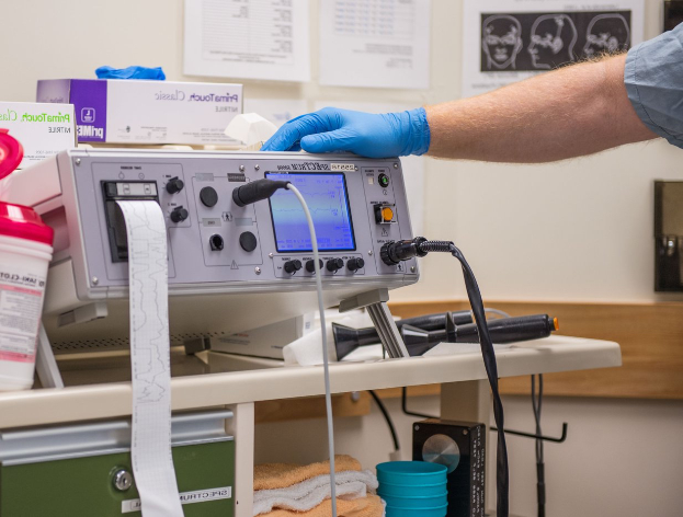 Eine Person in blauen Handschuhen arbeitet an einem medizinischen Gerät auf einem Tisch, der mit verschiedenen Gegenständen übersät ist, während im Hintergrund Papiere an der Wand hängen, was auf eine Krankenhausatmosphäre hindeutet.