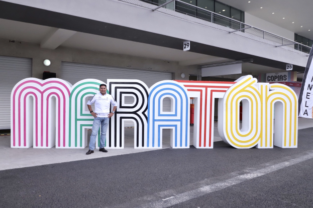 Ein Mann steht vor einem großen "Marathon"-Schild mit einem Banner rechts daneben, im Hintergrund ein Gebäude mit Lichtern, Geländern und Fensterläden.