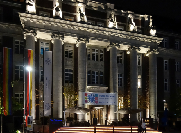 Großes neoklassizistisches Gebäude mit beleuchteten Säulen und Statuen, umgeben von Bäumen, Fahnen, Bannern, Treppen und Menschen, unter einem dunklen Himmel.