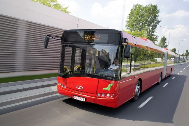 Roter Bus fährt auf einer Straße neben einem Gebäude mit einer Person im Inneren, neben einer Wand, Bäumen, Laternen und einem bewölkten Himmel im Hintergrund.
