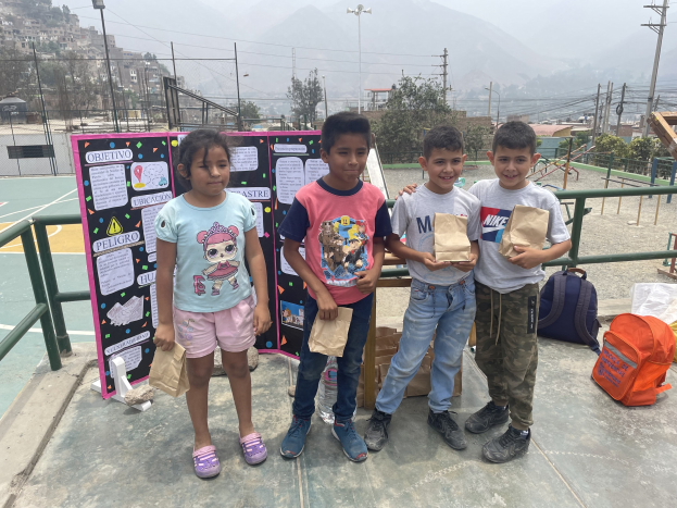 Gruppe von Kindern auf einem Basketballfeld mit Papiertüten in den H├Ąnden, mit Gegenst├Ąnden auf dem Boden rechts und einer Tafel mit Text links, vor dem Hintergrund von Z├Ąunen, Pfosten, Stra├čenlaternen, B├Ąumen, Geb├Ąuden, H├Ąngen und Himmel.