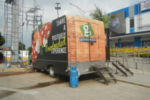 Ein großer Lastwagen steht vor einem Gebäude mit einer Tafel, auf der "Teilen Sie eine positiv überladene Erfahrung" steht, mit Stufen, einem Zylinder auf dem Boden, Geländern, Strommasten mit Drähten, Laternenpfählen, Bäumen und einem bewölkten Himmel.