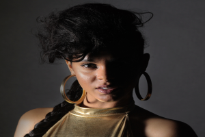 Eine Frau in einem goldenen Kleid und großen Ohrringen steht selbstbewusst vor einer Wand, ihre Haare sind glatt geschnitten und sorgfältig frisiert, sie trägt dezentes Make-up mit rosa Rouge und glänzenden Lippen und schaut direkt in die Kamera mit einem entschlossenen Gesichtsausdruck.