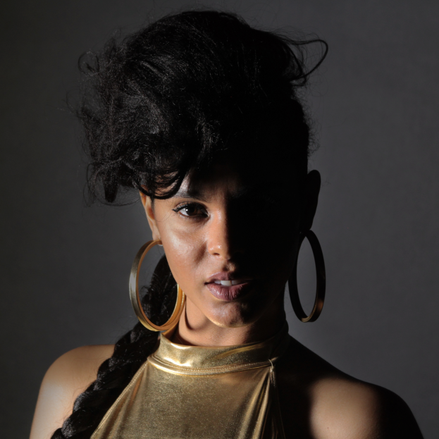 Eine Frau in einem goldenen Kleid und großen Ohrringen steht selbstbewusst vor einer Wand, ihre Haare sind glatt geschnitten und sorgfältig frisiert, sie trägt dezentes Make-up mit rosa Rouge und glänzenden Lippen und schaut direkt in die Kamera mit einem entschlossenen Gesichtsausdruck.