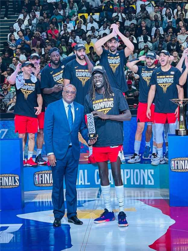 Eine Gruppe von Männern auf einem Basketballfeld, die Trophäen halten, mit Zuschauern im Hintergrund und zwei Trophäen auf Podesten.