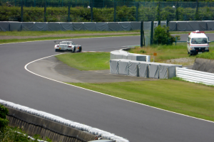 Ein Rennwagen fährt eine Rennstrecke entlang, im Hintergrund ein Polizeiwagen, umgeben von Gras, Pflanzen, Bäumen und Absperrungen, mit einem Fahrzeug auf der rechten Seite und Text unten.