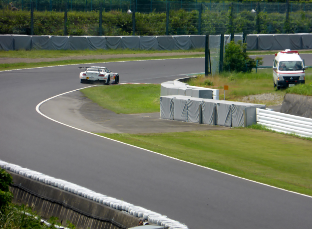 Ein Rennwagen fährt eine Rennstrecke entlang, im Hintergrund ein Polizeiwagen, umgeben von Gras, Pflanzen, Bäumen und Absperrungen, mit einem Fahrzeug auf der rechten Seite und Text unten.