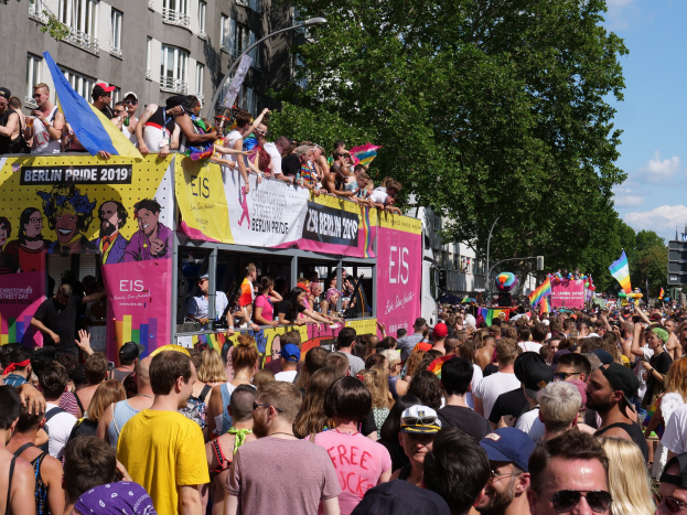 Eine große Menschenmenge marschiert auf der Straße bei der Berlin Pride 2019, viele tragen Mützen und Schutzbrillen, halten Fahnen, mit Bannern und Gebäuden im Hintergrund unter einem bewölkten Himmel.