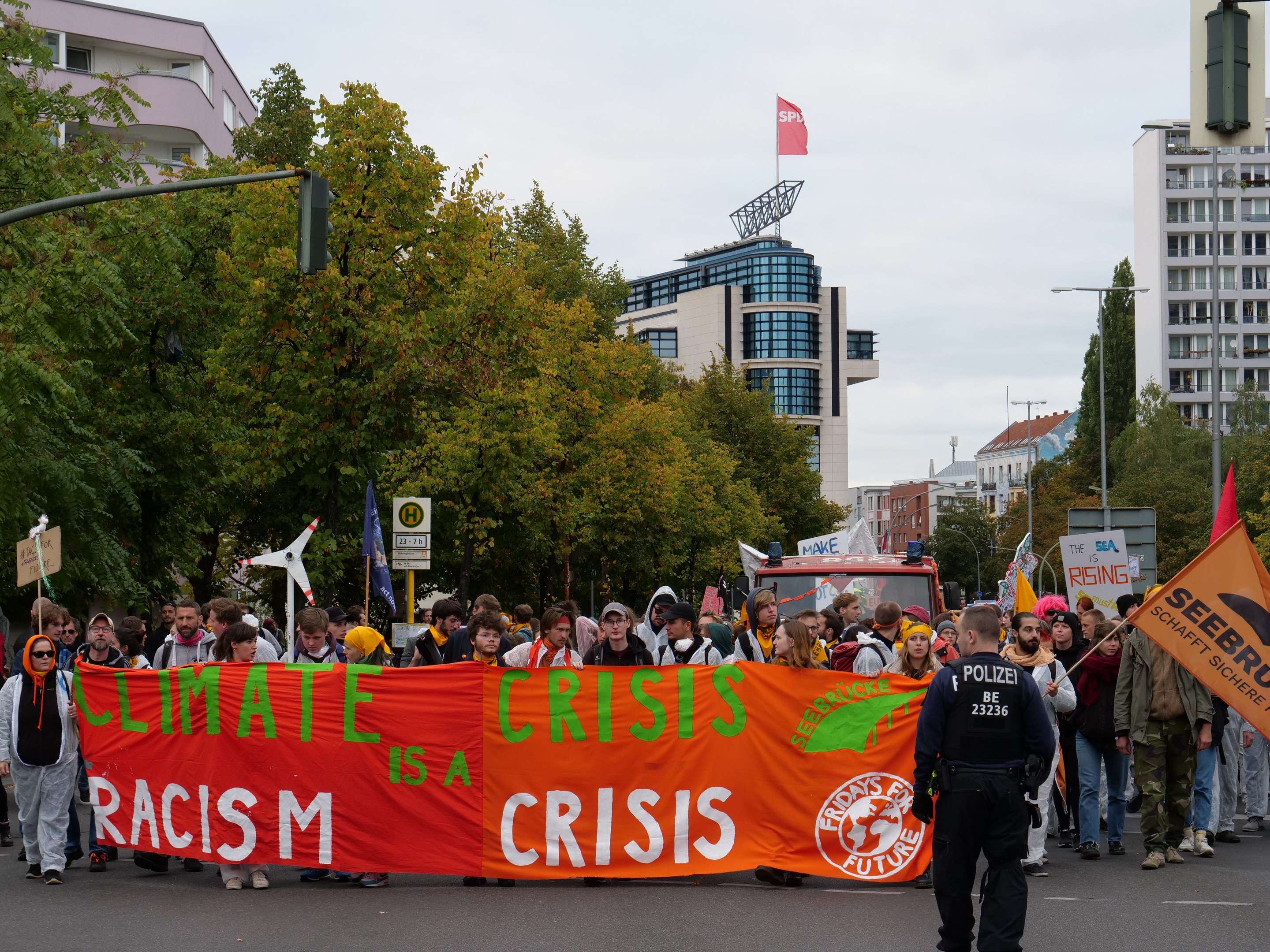 Eine Gruppe von Menschen marschiert auf einer Straße, die ein Banner mit der Aufschrift "Klima-Krise ist eine Krise" trägt, mit Bäumen, Laternenmasten und Schildern an der Straße, einem geparkten Fahrzeug in der Nähe und Gebäuden im Hintergrund unter einem klaren blauen Himmel.