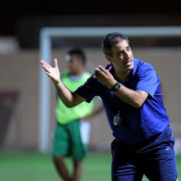 Ein Mann in einem blauen T-Shirt und Hose steht auf einem Fußballfeld und hält ein Mikrofon in der Hand, während er auf Spieler zeigt; im Hintergrund ist eine weitere Person in einem grünen Kleid und ein Torpfosten zu sehen.