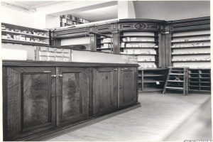 Ein Schwarz-Weiß-Foto eines Apothekeninneren mit einer Theke, Schränken, einer Leiter und Regalen voller verschiedener Gegenstände, mit Text oben drauf.