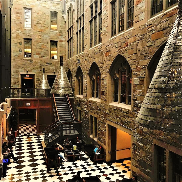 Großer Raum mit einem schwarz-weißen Schachbrettboden, einer zentralen Treppe mit Geländern, Fenstern, Deckenlampen und mehreren Personen an Tischen sitzend, die scheinbar eine Harry-Potter-Studio-Tour-Ausstellung in London besuchen.