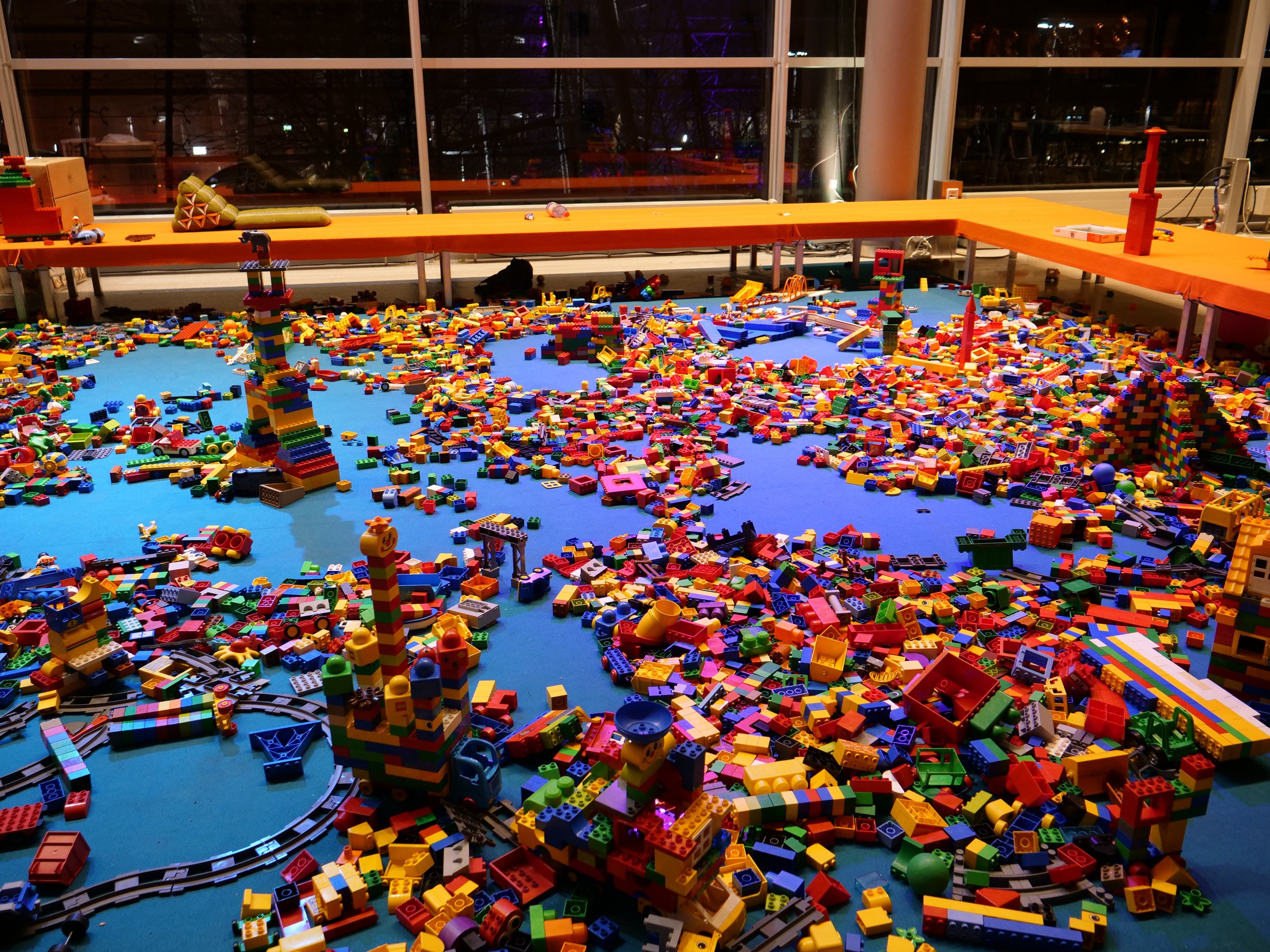 Ein großer Raum mit farbigen Bauklötzen auf dem Boden, umgeben von Tischen und Säulen, mit einem Blick durch Glasfenster auf Menschen, Lichter und Ausstellungsstücke in einem Lego-Museum.