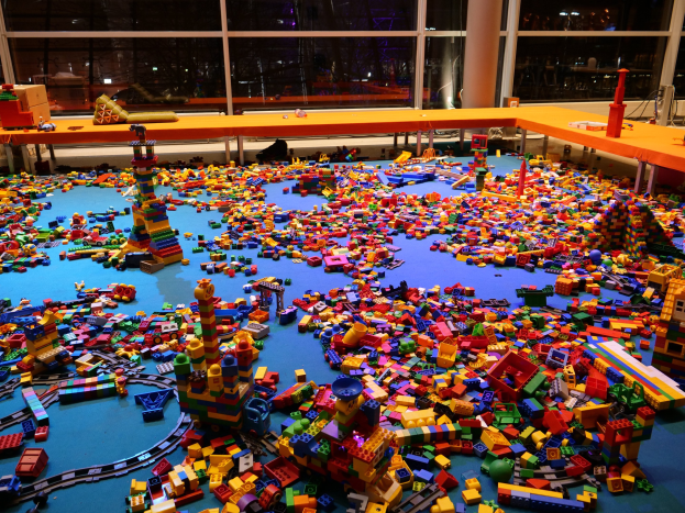 Ein großer Raum mit farbigen Bauklötzen auf dem Boden, umgeben von Tischen und Säulen, mit einem Blick durch Glasfenster auf Menschen, Lichter und Ausstellungsstücke in einem Lego-Museum.