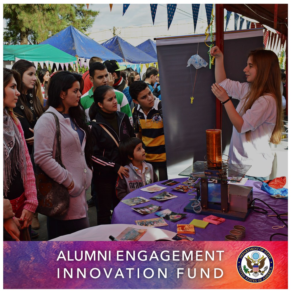 Gruppe von Menschen um einen Tisch versammelt mit einer "Alumni Engagement Innovation Fund"-Aufschrift, umgeben von Gegenständen, mit Zelten und Bäumen im Hintergrund unter einem klaren blauen Himmel.