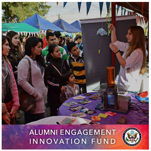 Gruppe von Menschen um einen Tisch versammelt mit einer "Alumni Engagement Innovation Fund"-Aufschrift, umgeben von Gegenständen, mit Zelten und Bäumen im Hintergrund unter einem klaren blauen Himmel.