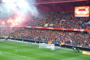 Ein Fußballspiel in einem großen Stadion mit einem Tor, Fans auf dem Rasen, Bannern, Fahnen, einem Display, Metallrahmen, Deckenleuchten und Rauch, der vom Feld aufsteigt.