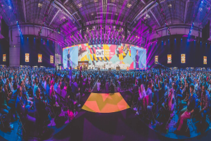 Eine große Menschenmenge sitzt auf Stühlen, die einer hell erleuchteten Bühne zugewandt sind, auf der mehrere Personen stehen und von Bildschirmen umgeben sind, wahrscheinlich auf einer Online-Marketing-Rockstars-Veranstaltung.