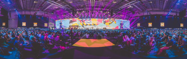 Eine große Menschenmenge sitzt auf Stühlen, die einer hell erleuchteten Bühne zugewandt sind, auf der mehrere Personen stehen und von Bildschirmen umgeben sind, wahrscheinlich auf einer Online-Marketing-Rockstars-Veranstaltung.