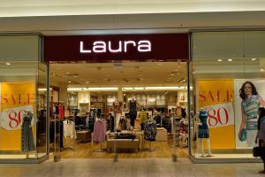 Ein Laura Modegeschäft in einem Einkaufszentrum mit Schaufensterpuppen in Kleidern, hängenden Kleidern, Informationsschildern, Deckenbeleuchtung und einer Person, die mit einer Tasche in der Hand geht.