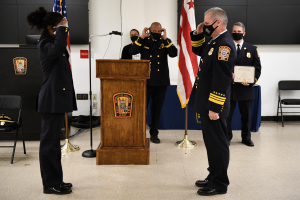 Gruppe uniformierter Personen in Masken vor einem Podium mit einem Mikrofon während einer Vereidigung eines neuen Feuerwehrchefs, der eine Urkunde hält und von Fahnen, Stühlen, Tischen und einer Wand umgeben ist.