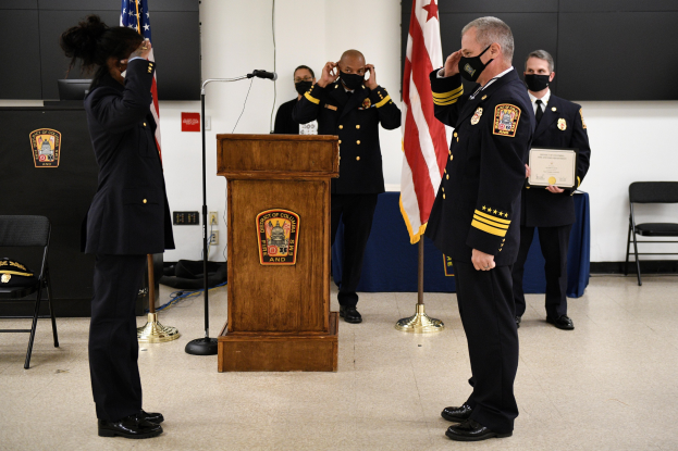 Gruppe uniformierter Personen in Masken vor einem Podium mit einem Mikrofon während einer Vereidigung eines neuen Feuerwehrchefs, der eine Urkunde hält und von Fahnen, Stühlen, Tischen und einer Wand umgeben ist.