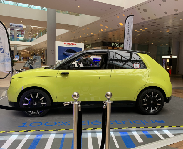 Gelber Wasserstoff-elektrischer Auto vor einem Gebäude mit Absperrpfosten, Bannern, Menschen, Schildern, einer Topfpflanze, Deckenleuchten und Lüfter.