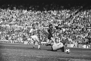 Schwarzes Foto von Männern, die auf einem Feld Fußball spielen, mit Zuschauern im Hintergrund und einer "Bundesliga 1978-79 Ajax 1-1 Hannover 96"-Fahne.