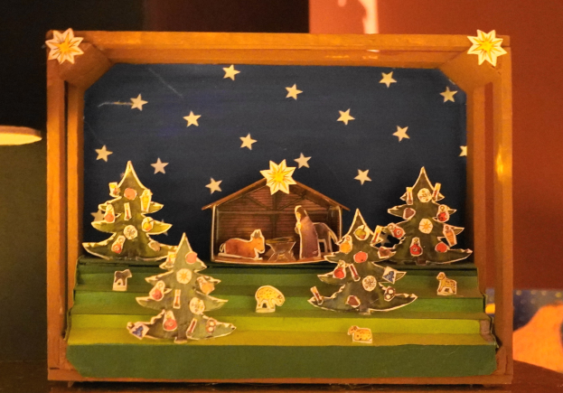 Eine Krippe des Weihnachtsfestes in einer Holzkiste auf einem Tisch, mit einer Lampe links und einer Wand im Hintergrund, mit Dekorationen wie Weihnachtsbäumen, Tieren und einem Haus.