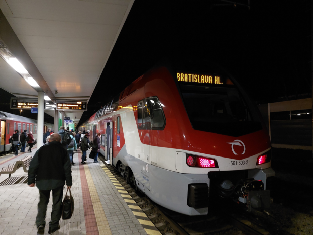 Roter und weißer Zug an einem nächtlichen Bahnhof mit Passagieren auf dem Bahnsteig und Bänken auf der linken Seite unter Deckenbeleuchtung.