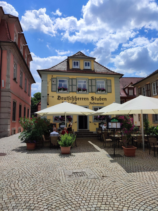 Außensitzbereich mit Tischen, Sonnenschirmen und Menschen vor einem gelben Gebäude mit der Aufschrift "Deutschen Stuben Restaurant" in Baden-Württemberg, Deutschland, unter einem blauen und weißen Himmel.