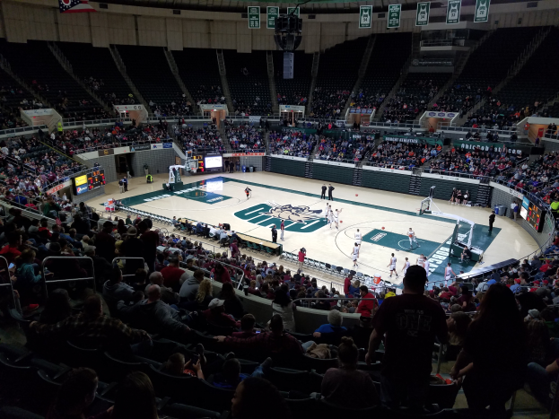Eine große Arena voller Zuschauer bei einem Basketballspiel, mit einem Spielfeld in der Mitte und Anzeigetafeln, Scheinwerfern und anderen Gegenständen oben.
