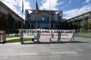 Gruppe von Menschen mit einem Banner, auf dem "Deutschland, Lagerland Migration ist kein Verbrechen" steht, vor einem Gebäude mit Fenstern, umgeben von Gras, einem Metallzaun, Pflanzen, Bäumen und Fahnen auf Stangen, unter einem bewölkten Himmel.