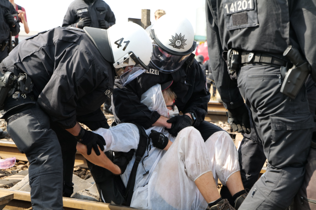 Eine Gruppe von Polizisten in Einsatzausrüstung verhaftet einen Mann in einem weißen Kleid auf einem Bahngleis, mit anderen im Hintergrund.